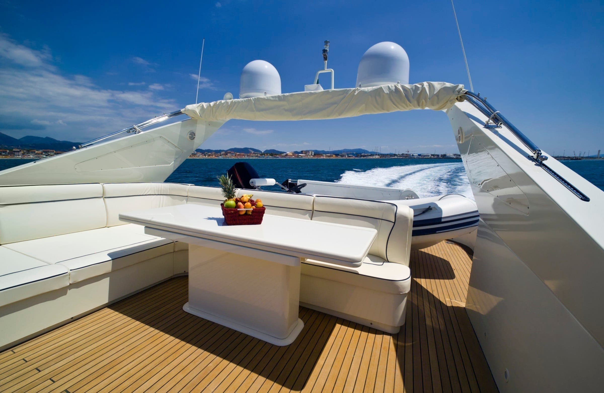 The upper deck of a yacht at sea, a white banquette and a table laid with fruit, the coast in the distance.