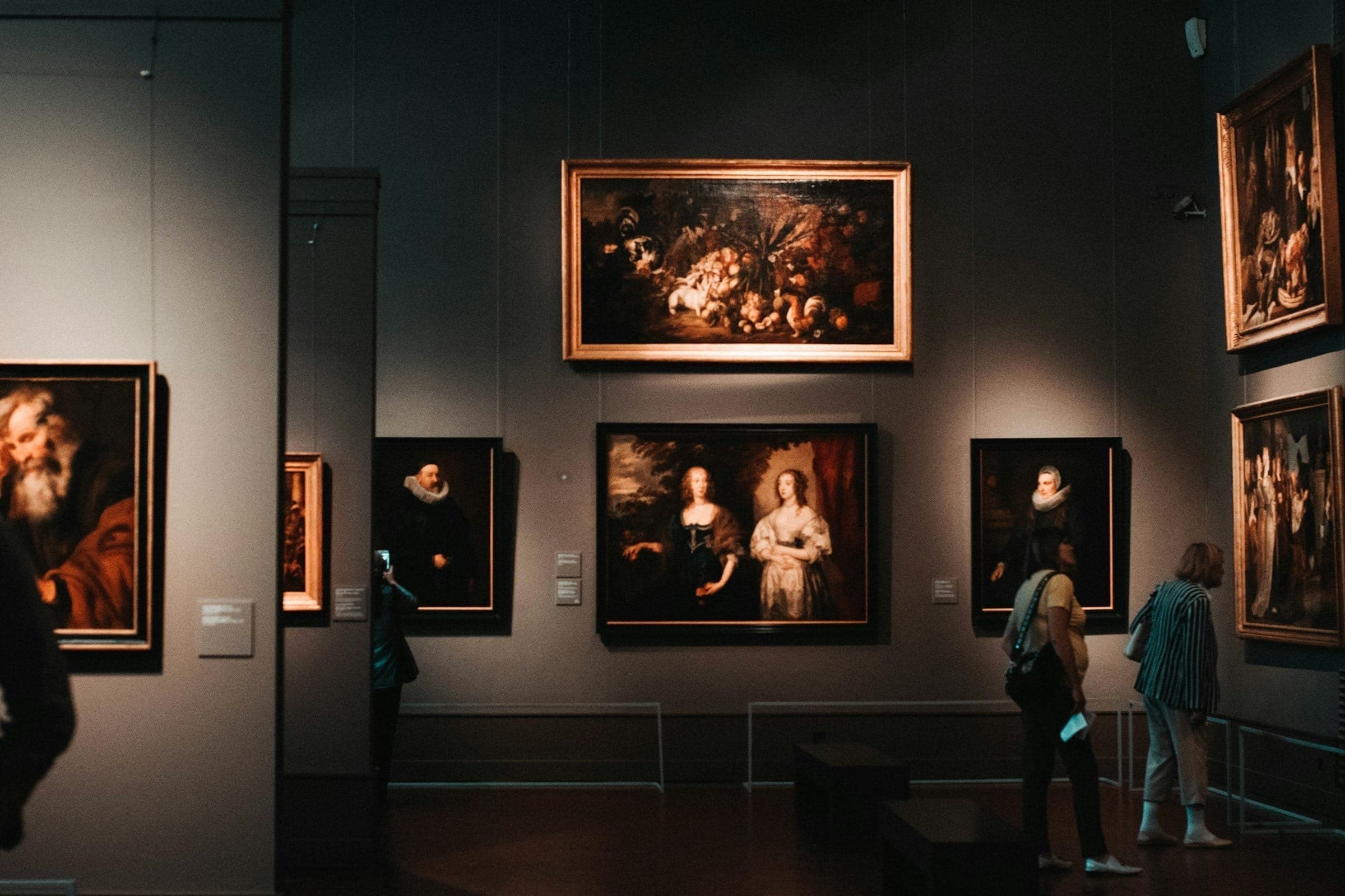 A museum gallery hung with gilt-framed paintings, a few visitors in front of the works.