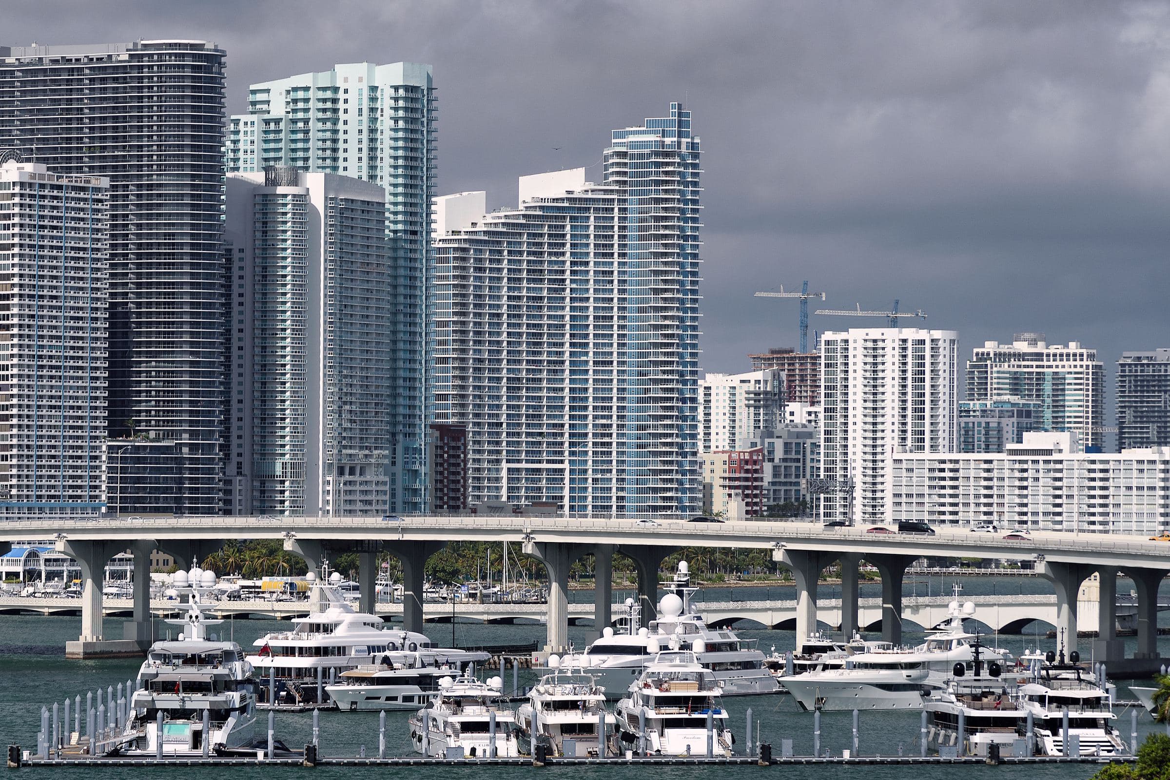 Biscayne Bay: yachts moored beneath a bridge, Miami towers under a stormy sky.