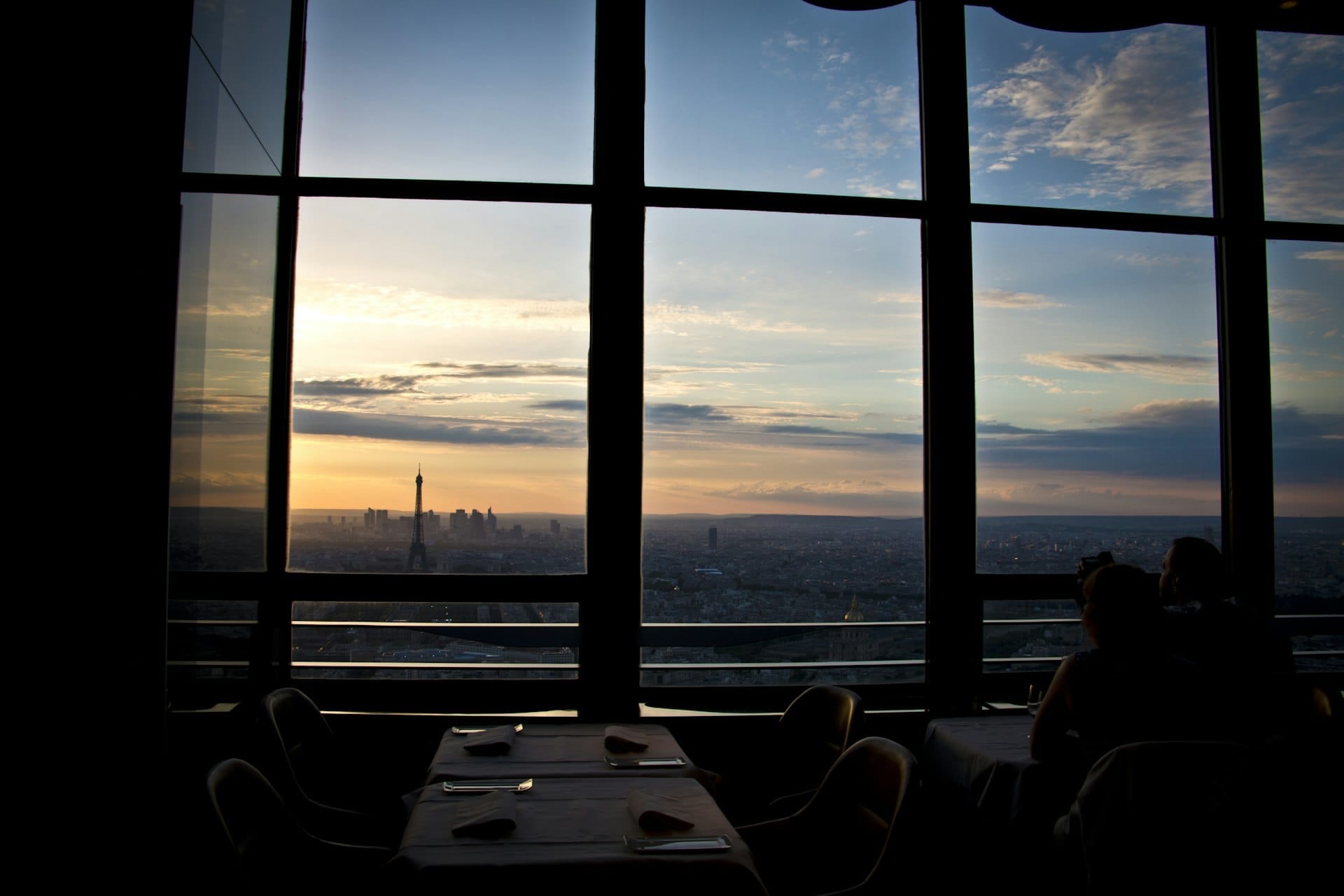 Une salle sombre au crépuscule, les tables dressées, Paris derrière la baie vitrée.