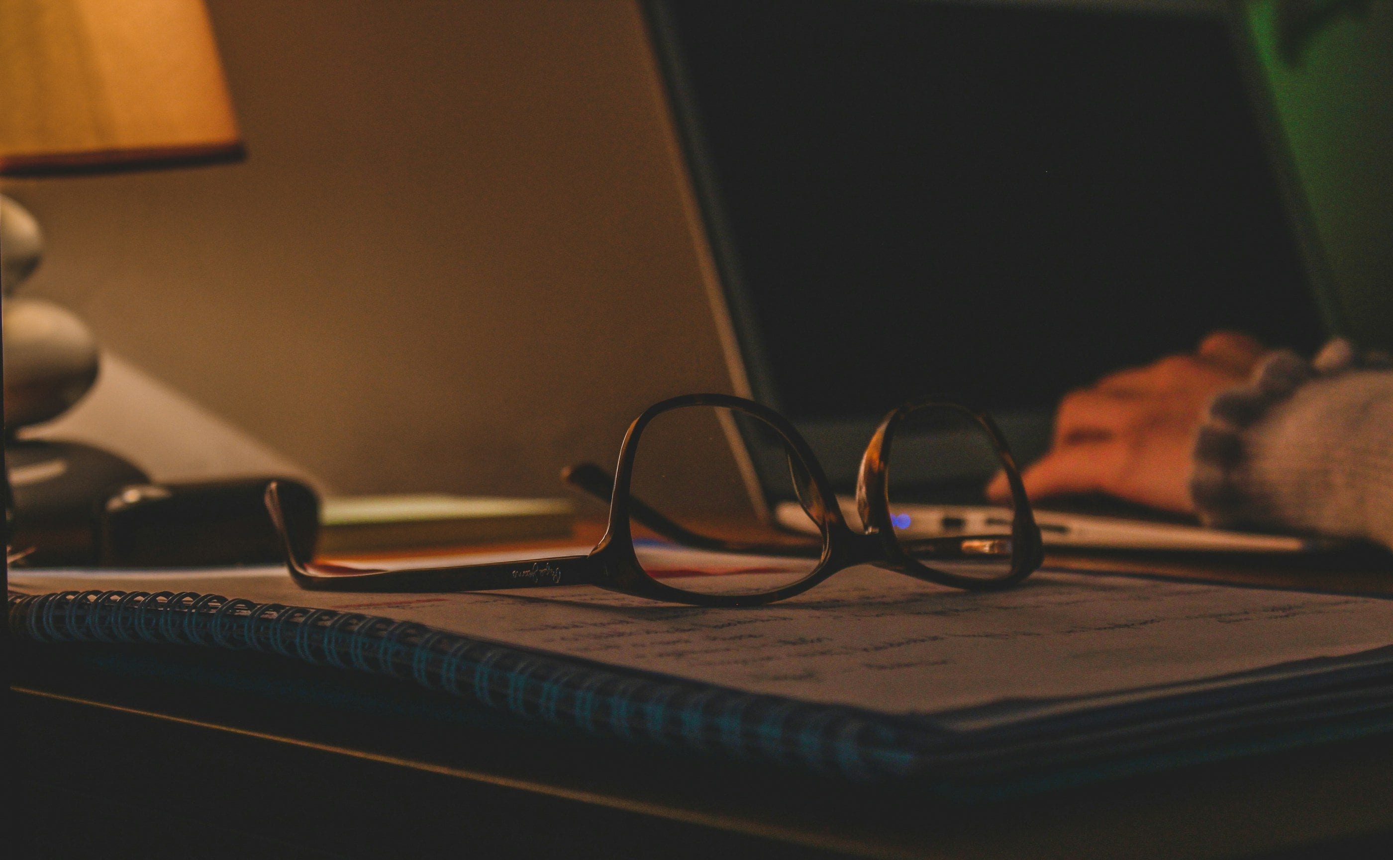 Reading glasses resting on a notebook late in the evening, beside a lit lamp.