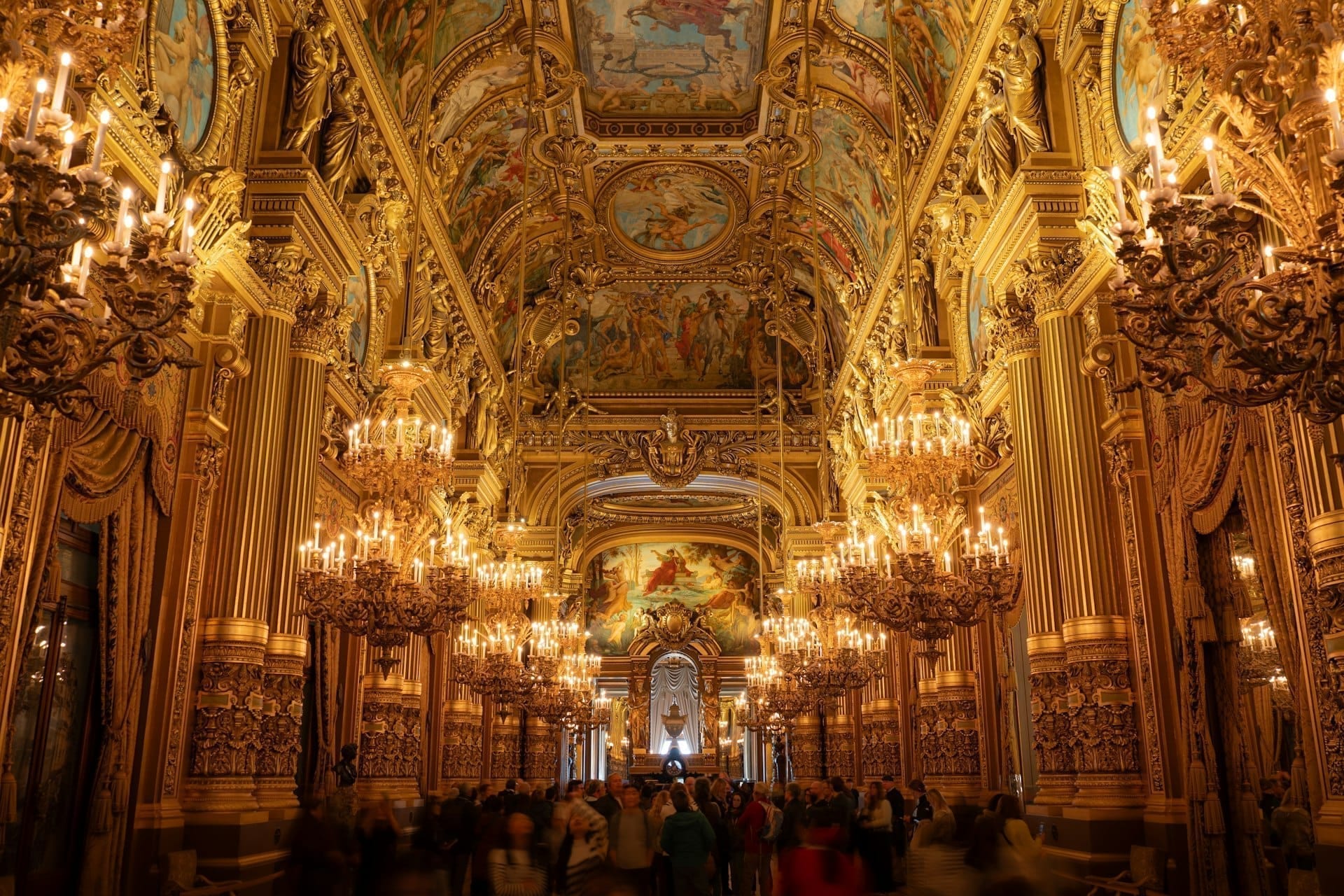 Un grand foyer doré d'opéra, plafonds peints et lustres de cristal allumés.