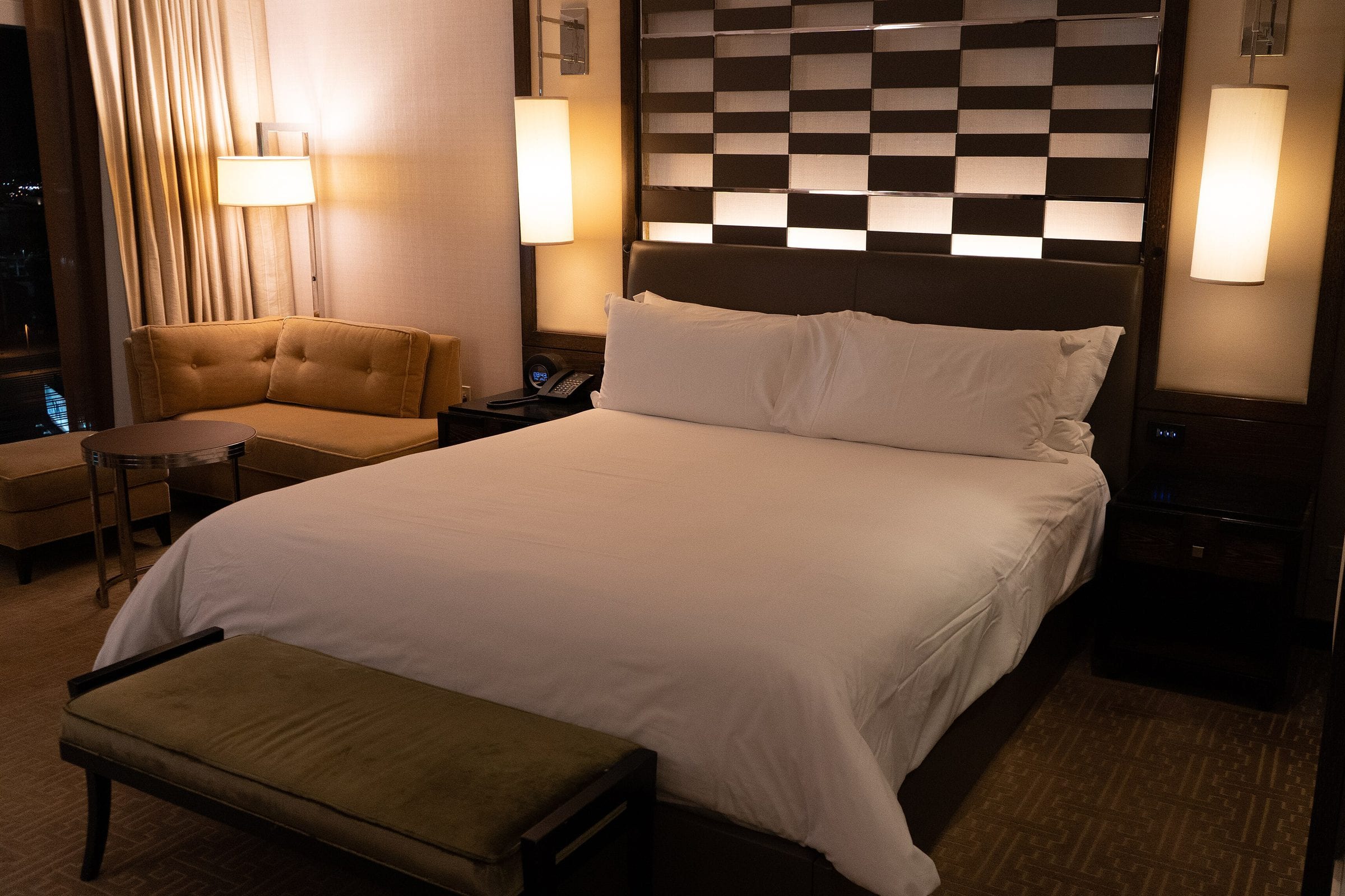 A hotel room at night, bed made up in white and the city beyond the window.