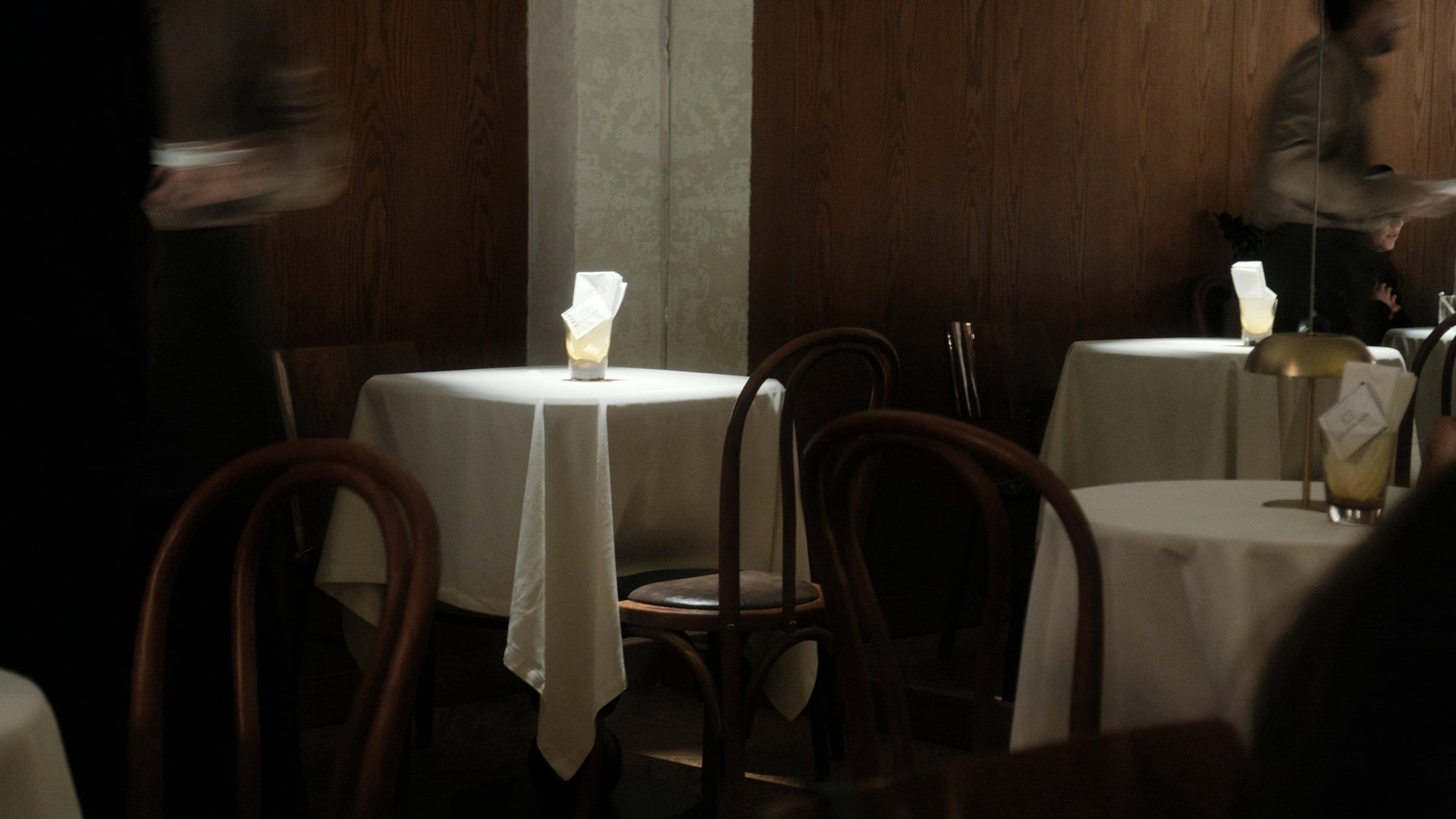 Un comedor en servicio, de noche: manteles claros, lámparas de mesa, un camarero en movimiento.