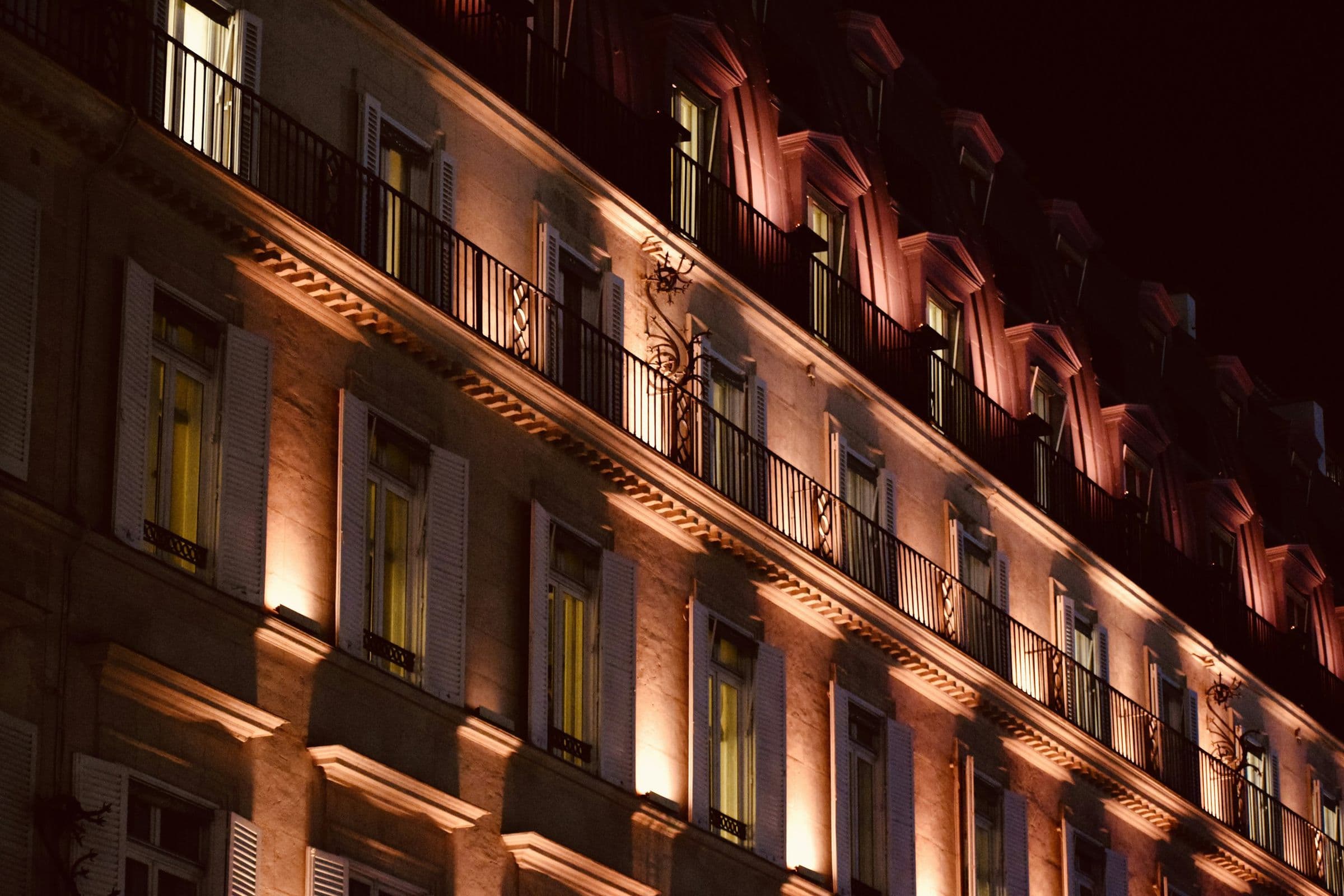 The façade of a grand hotel lit up at night, wrought-iron balconies and pale shutters.