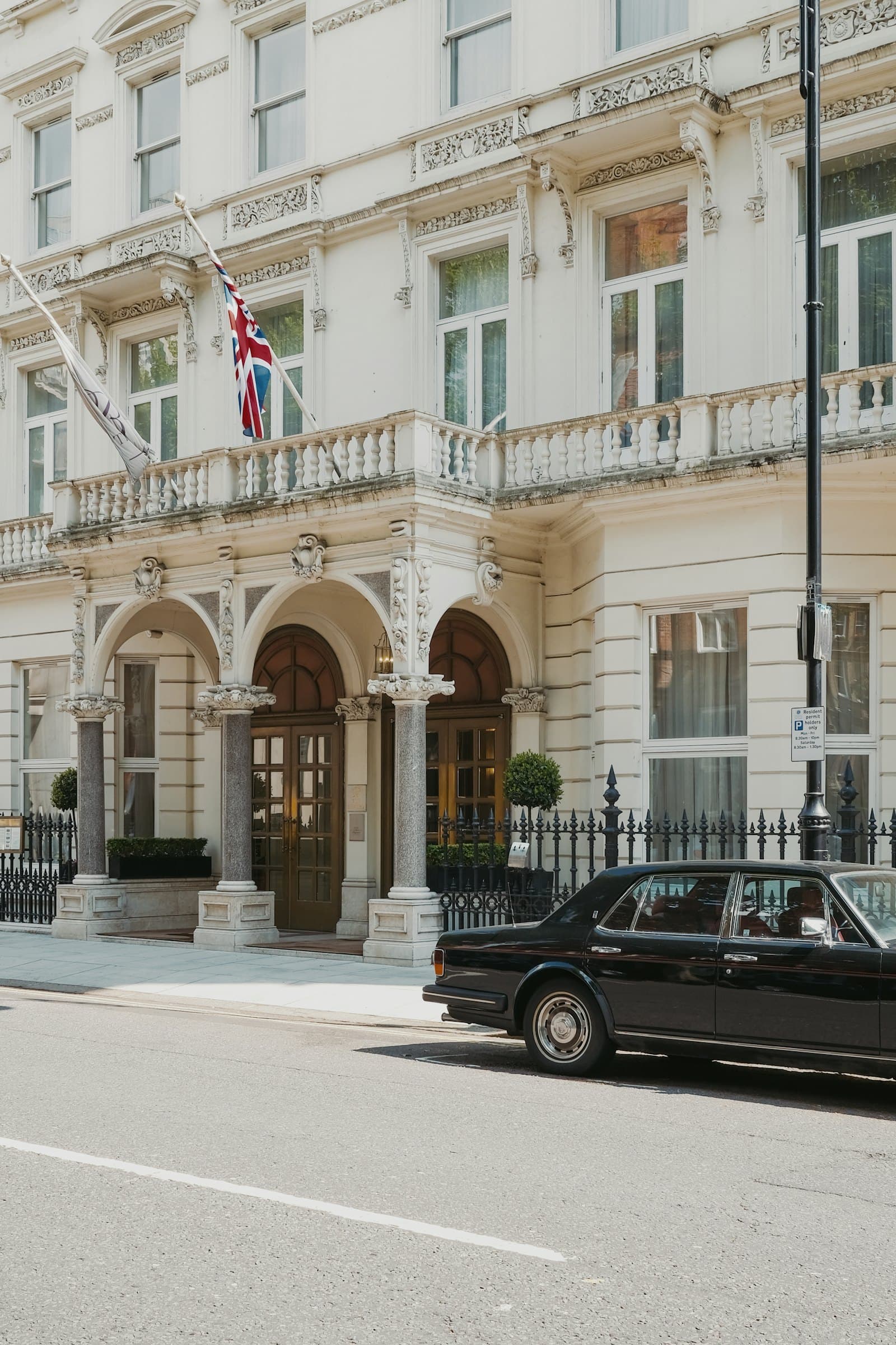 A black car waiting before a white stucco portico, in London, in summer.
